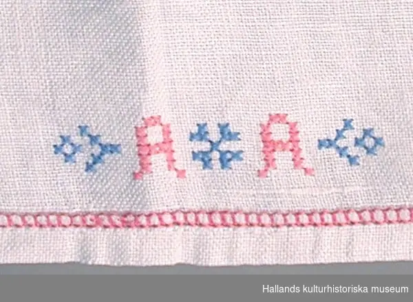 Duk av tuskaftat linne, broderad med korsstygn i rosa och blått samt utdragssöm sydd med rosa garn. Märkt: "AA 1943".