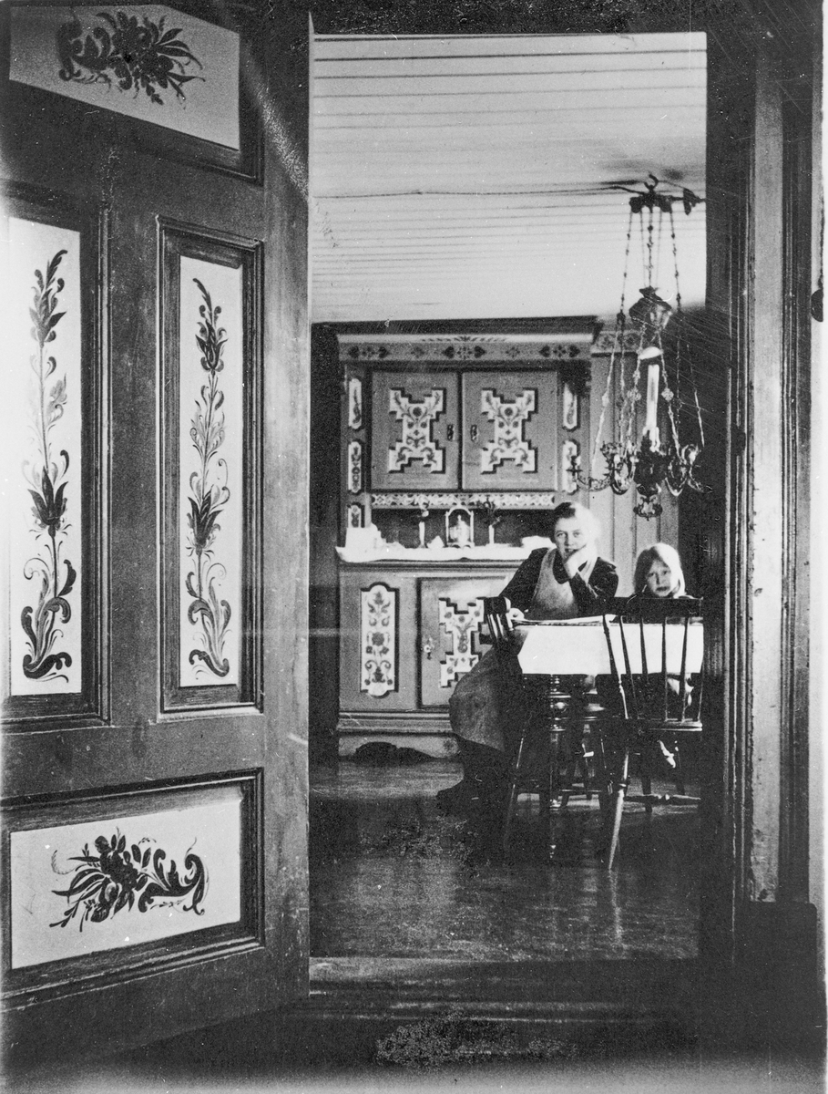 Interiør fra gamlebygningen på Nedre Kopseng, ca. 1920. Liv Nyhus (f. Blegeberg) og Anna Juvet (f. Blegeberg) ved bordet. Interiøret er malt av Karl Lunde.