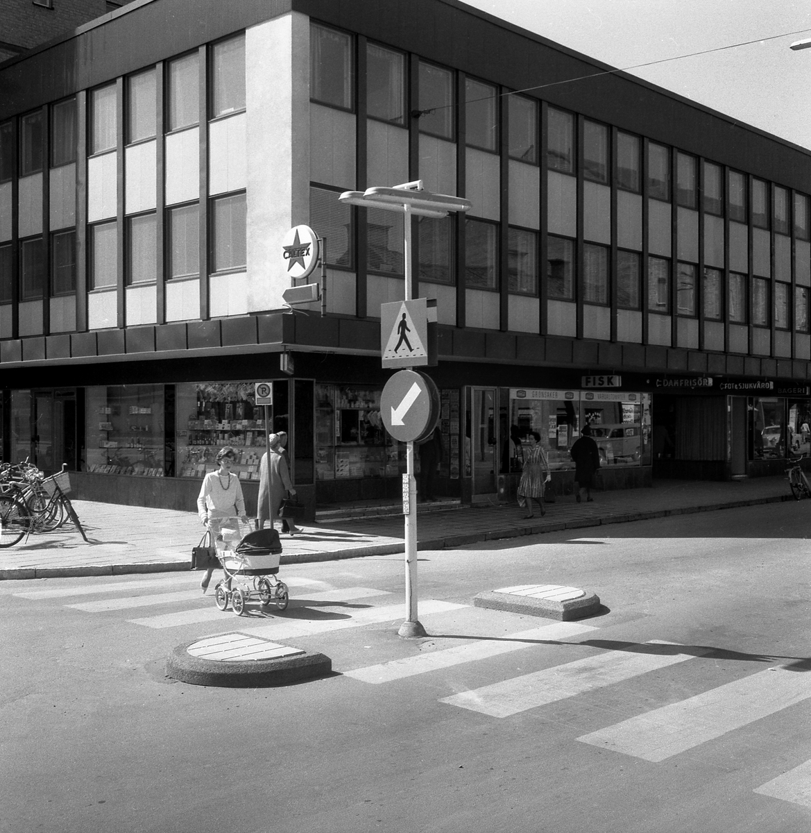 Motiv från Linköping 1964. Bildens egentlinga syfte var sannolikt att  uppmärksamma innovationen med refuger vid övergångsställen. Vi betraktare av senare tid får en glimt av miljön av Storgatans nedre del där gångstråkets placering förklaras av att biltrafik ännu var tillåten från och till Hamngatan.