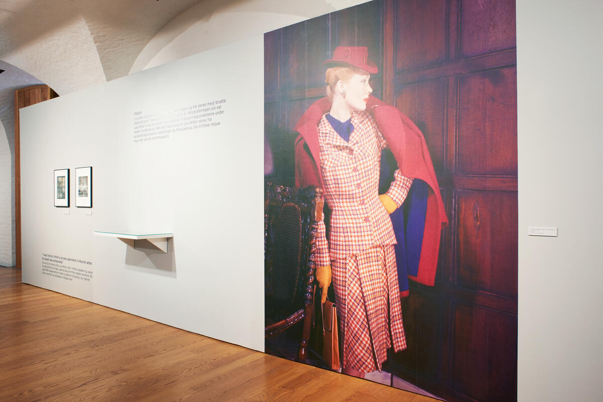 Exhibition room with a large picture of a female model dressed in a checkered skirt and jacket. She has a red coat over her shoulders and a red hat on her head.