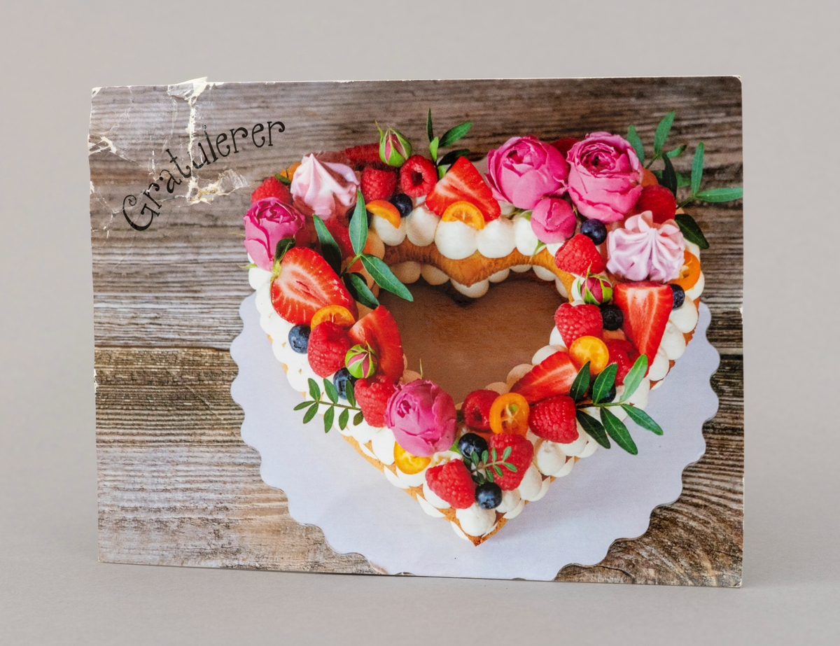 Gratulasjonskort med motiv av hjerteformet kringle, pyntet med blomster, marengs og frukt på forsida.