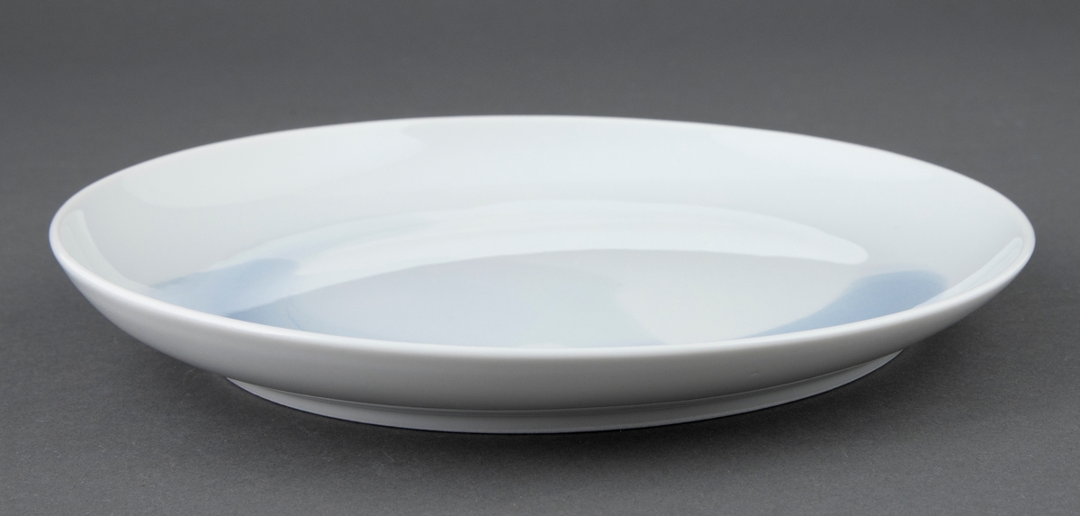 Rund tallerken i hvit porselen med utflytende blå underglasur i bunnen. Svak helling på fatet slik at det er høyere på den ene siden.