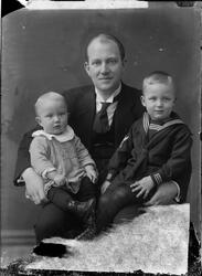 Portrett av mann med to barn. Barnet til venstre er kledd i 