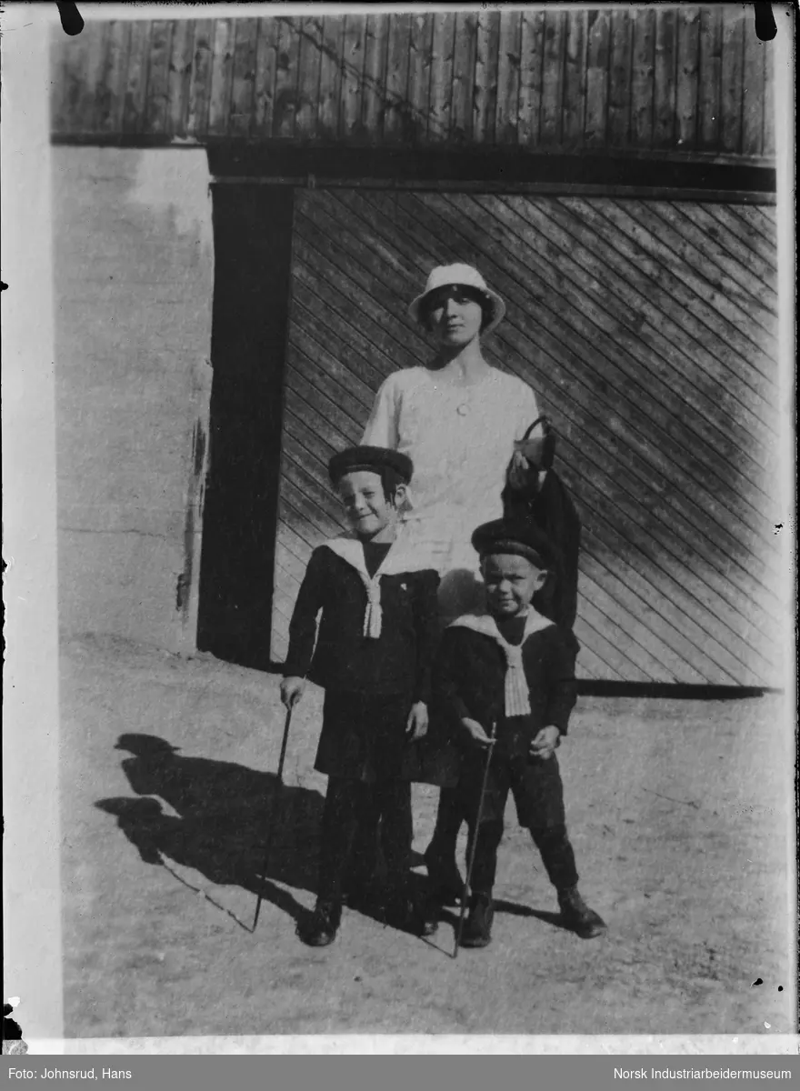 Familieportrett. En kvinne og to gutter stående foran port. Kvinnen i kjole og guttene i matrosdress med pinne i hånden.