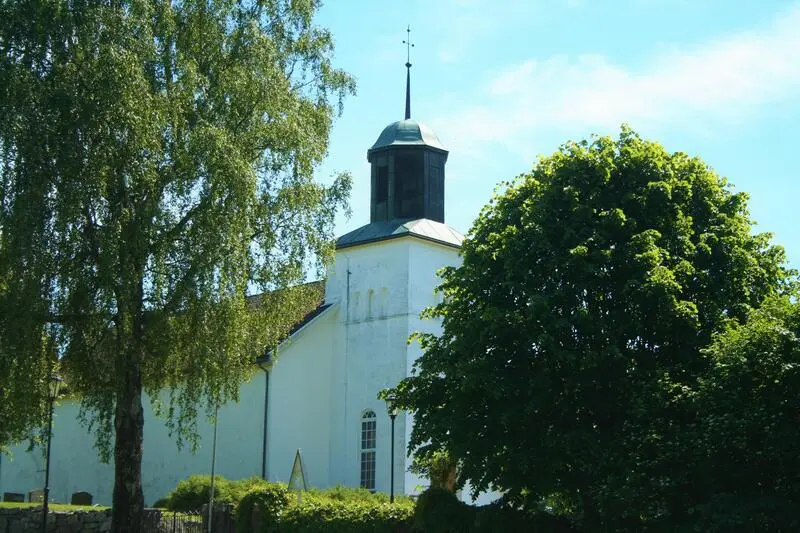 Bildet viser en hvit kirke mellom grønne trær. Det er sol og sommer. Dette er Botne kirke.