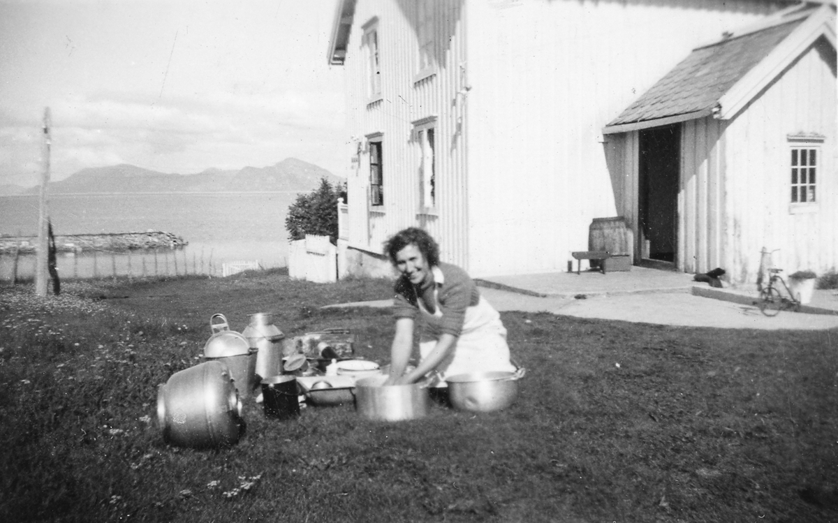 Kvinne vasker opp på plenen foran et hus. En molo og fjell i bakgrunnen.