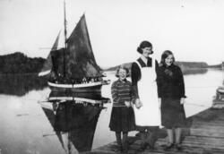 Kvinne og 2 jenter på kaia. Båt og mannskap på sjøen i bakgr