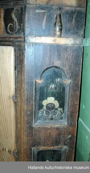 Hörnskåp av brunbetsat trä. Nerfällda speglar på väggarna med blommor målade i brunt och vitt. På skåpets övre del målat årtalet 1777 och initialerna NLS. I skåpet tre hyllor och nederst en låda.