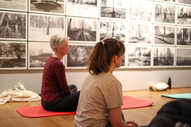 To kvinner sitter på yogamatter inne i et museumsrom.
