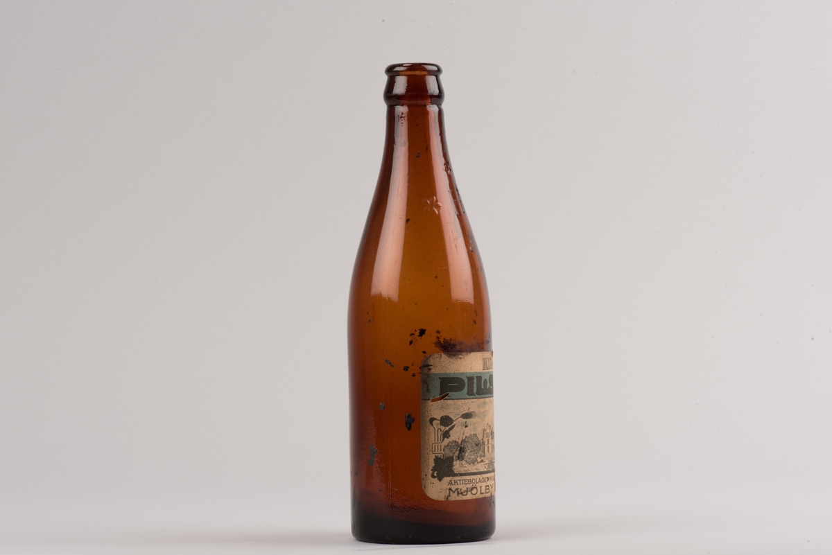 Pilsnerflaska i brunt glas.
På flaskan finns ett H omgivet av tre stiliserade blommor. En etikett med text och motiv visar att pilsnern var bryggd på Mjölby bryggeri. Motivet på etiketten är bryggeribyggnaden.