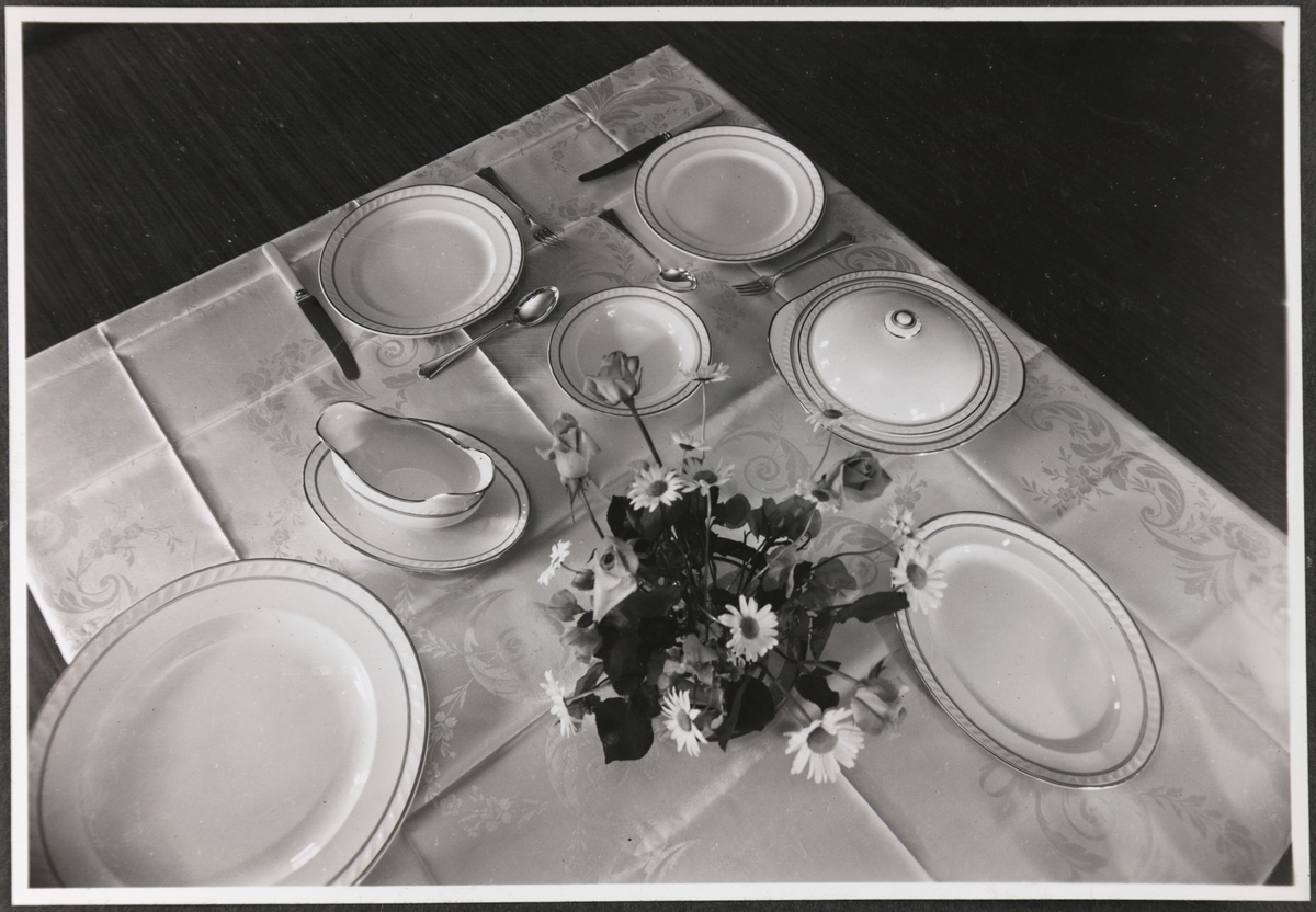Fotoalbum fra Stavangerflint A/S med produktbilder.
Fotografiet viser eit bord dekt med middagsserviset "Skaugum", med middagstallerknar, bestikk, serveringsfat og sausenebb, duk og blomster i vase. Formgiver til "Skaugum" var Thorbjørn Feyling.