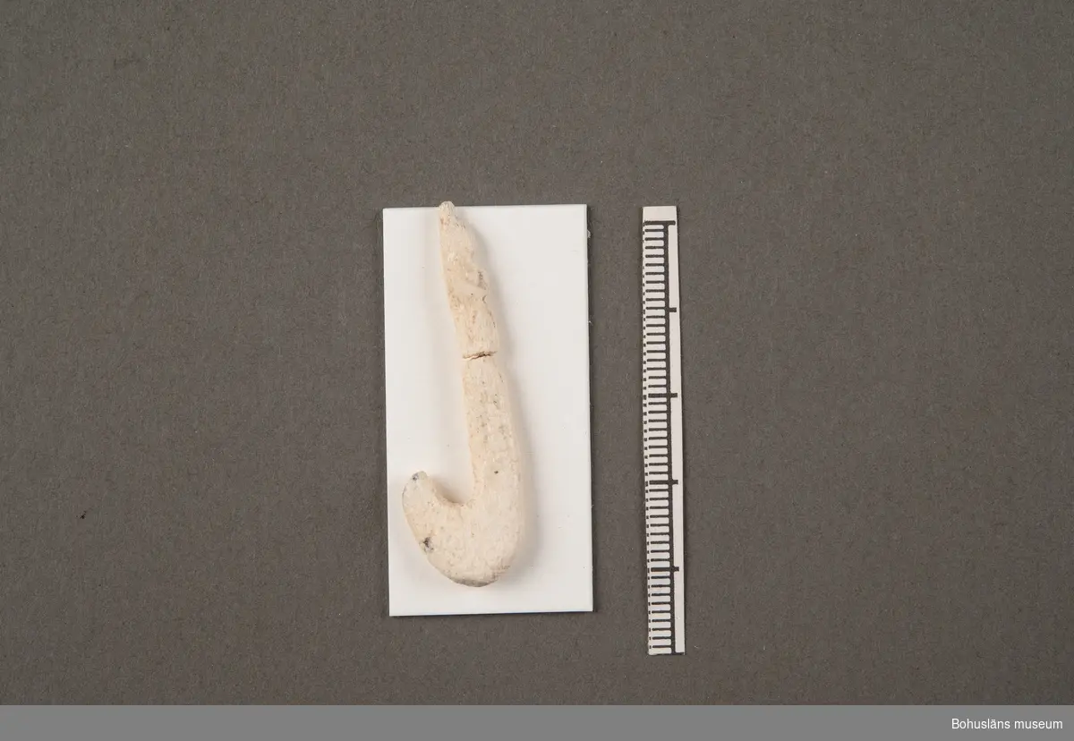 Fiskekrok av ben, relativt stor modell. Kroken är skadad, i två delar, men sammanfogad och fastmonterad på en bit vit papp. Saknar yttersta spets och hulling. Skaftdelen har 2 inhak för fäste av lina. Kroken är ca 8 000 - 9 000 år gammal.