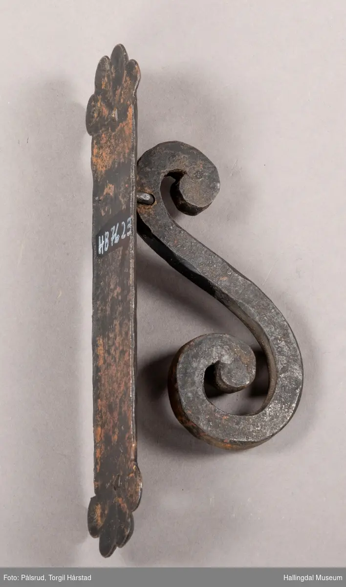 Dørhammer. En hammer i S-form som er hengslet høverst, estet på beslag som skrus fast i dør. 