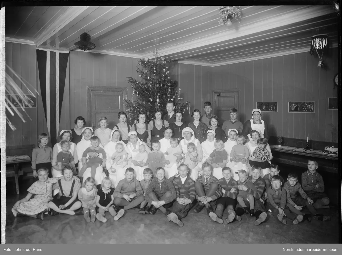 Julaften på barnehjemmet. Barn, ungdom og ansatte oppstilt i fellesrom med juletre i bakgrunnen.
