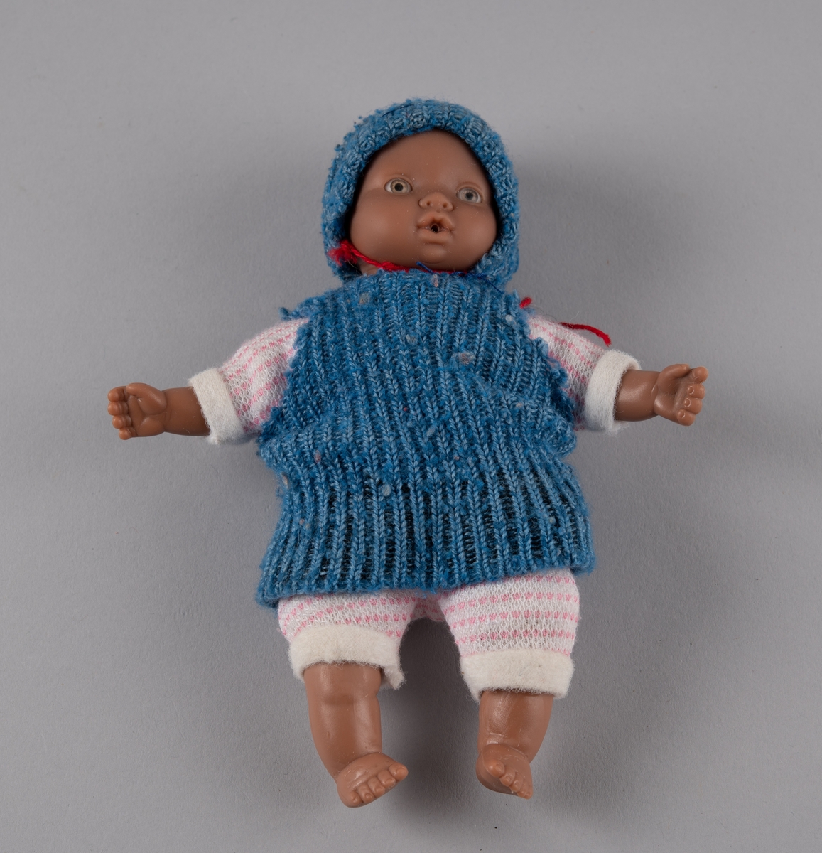 Babydukke med hode, armer og føtter av plast, og torso av tekstil fylt med vatt. Den er kledd i hjemmestrikket, rosa og hvit buksedrakt, samt blå genser og lue. Lua har en rød hakesnor.