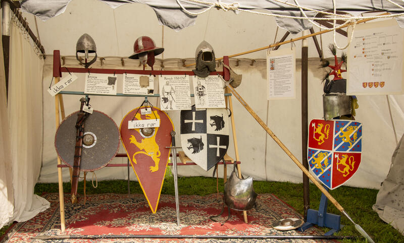 Utstilling med runde, trekantet og spisse skjold med ulike våpenskjold i rødt, blått, gult, hvitt og svart. Plakater med skrift om riddersystemet i Norge i middelalderen og metallhjelmer