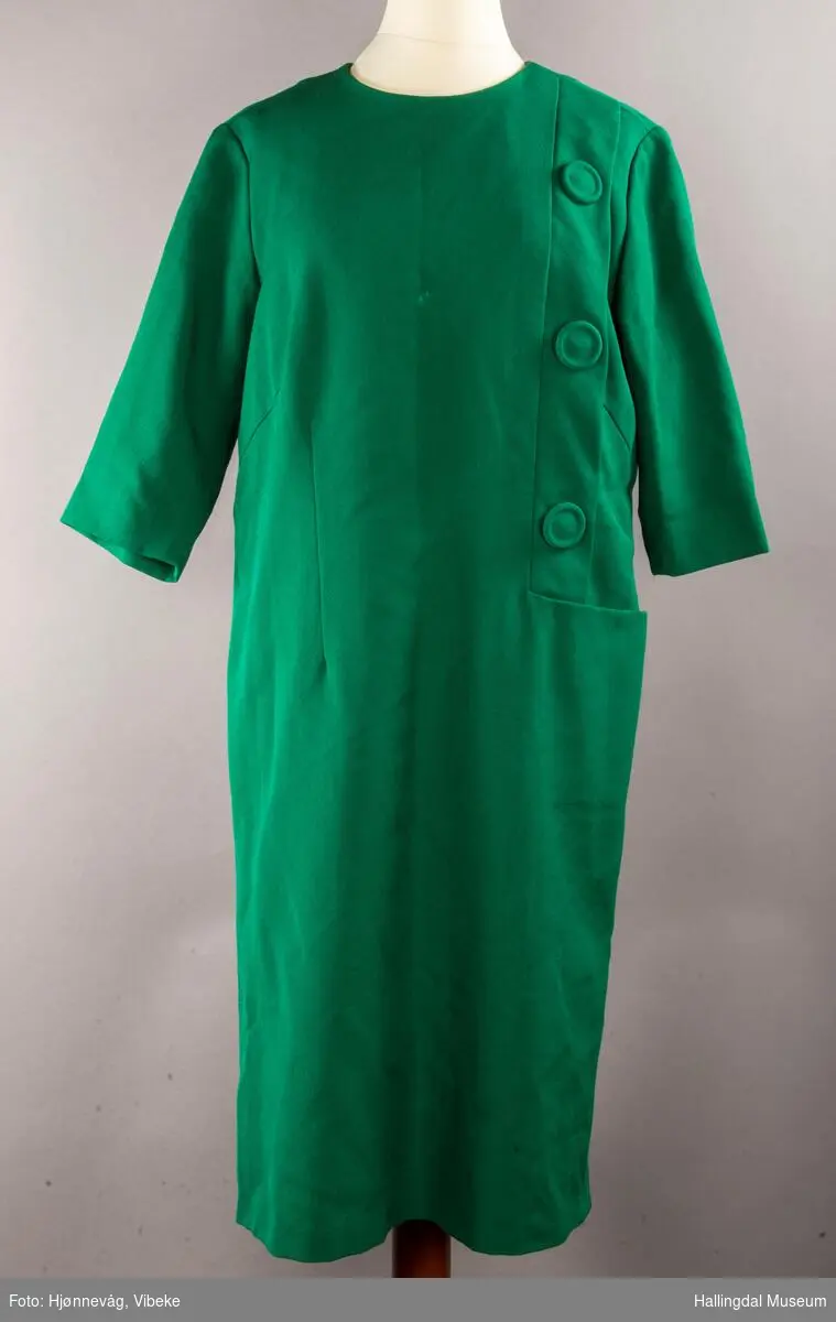 Grønn kjole med halvlange ermer. 3 store knapper på venstre side som er festet på en stolpe. Ei stor lomme under stolpen. Glidelås i ryggen. Rund hals.