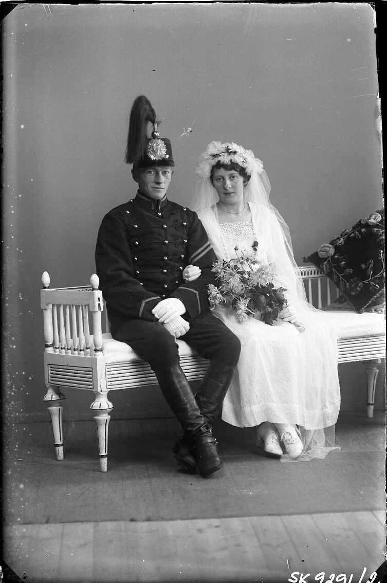 Bröllopsfotografi med Axel Gran och Ester Blom. De sitter i en låg soffa av gustavianskt snitt. Mannen bär militär uniform (vicekorpral?) med kask på huvudet. Kvinnan har slöja och en bukett i handen.