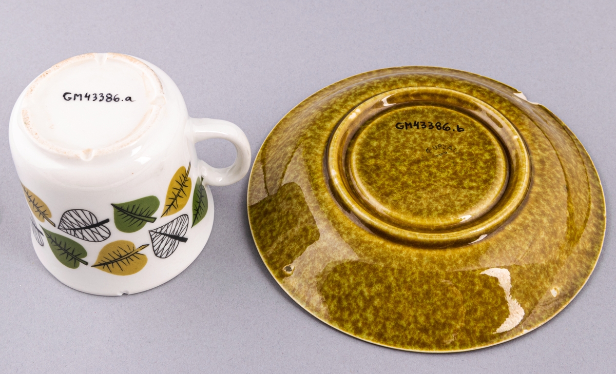 Kaffekopp med fat, i flintgods. Modell EF, dekor Bladranka. Vit glasyr med grönt bladmönster i grönt och gulbrunt, fatet gulbrunt. Skapad av Berit Ternell för Gefle Porslinsfabrik 1965.