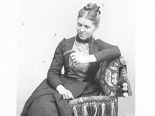 Fotograf Mathilda Ranch sitter på en stoppad stol med en liten fågel på handen. Stolen är av ottomansk typ med kraftiga fransar och en stor tofs med bollar baktill.
