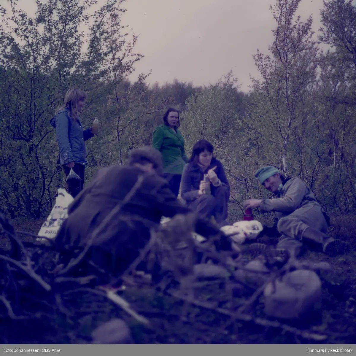 Foto av tenåringer på skogstur, antagelig bærplukkere i Finnmark. En mann ligger på bakken til høyre med en termos i hånden

Foto trolig tatt på 1970-tallet