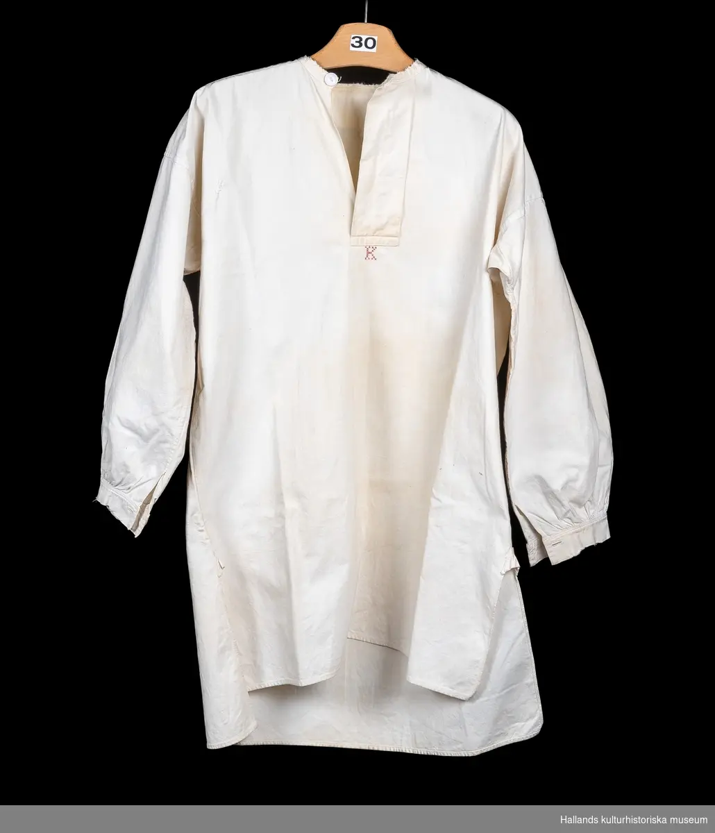 Skjorta, så kallad slåtterskjorta, använd vid slåtter. Av vit bomull. Bakstycket går något längre ner än framstycket. Långärmad med knapp vid manschetten. Sprund med knapp vid halsen. Rund hals. Märkt: "K", med korsstygn. En lagning. Lite sliten i kanterna.