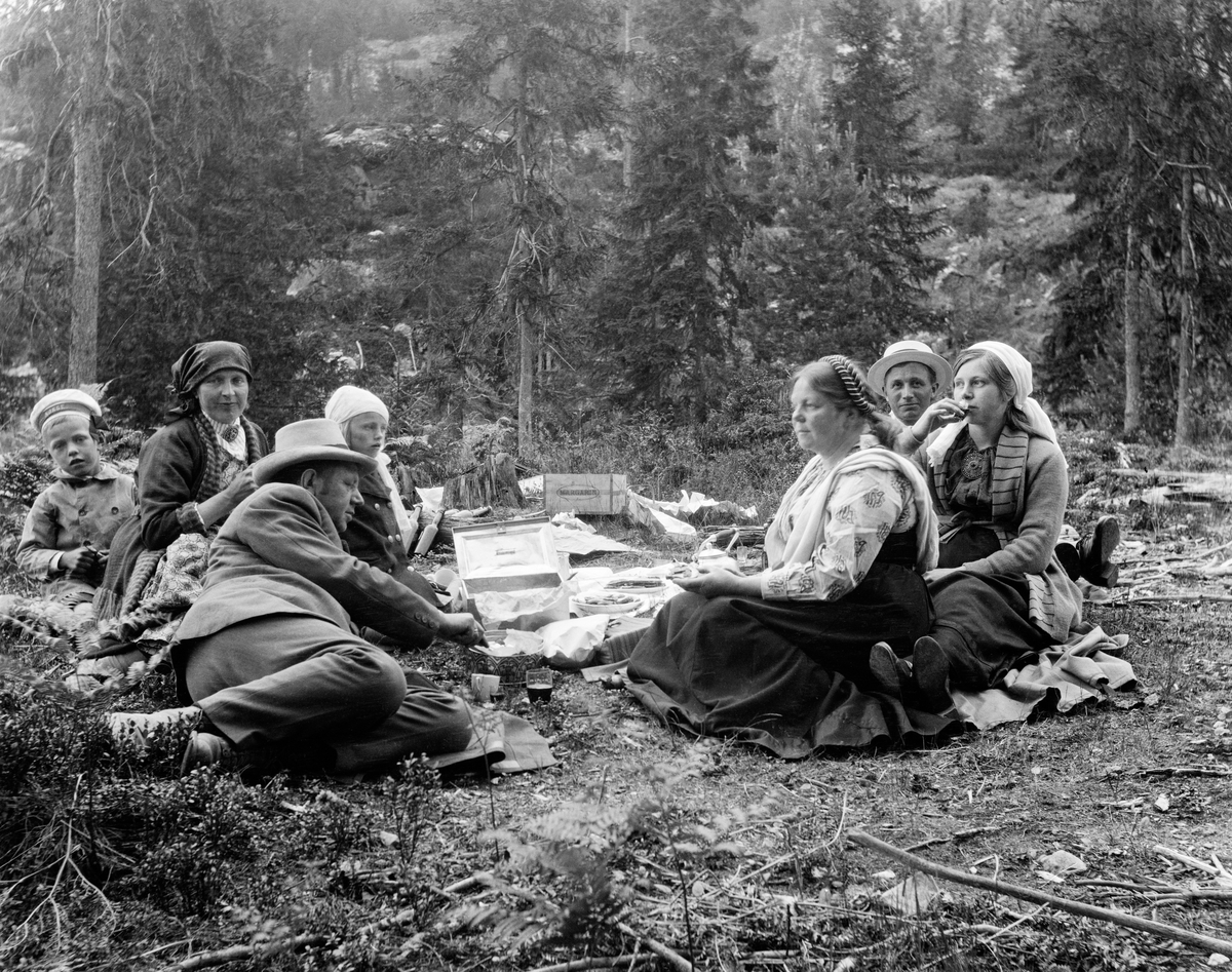 Bilde av en gruppe voksne og barn som trolig er familien Midttun og familien Folkestad på tur til Reskjem sommeren eller høsten 1923. De har satt seg ned på ei flate i skogen med medbragt mat og drikke.