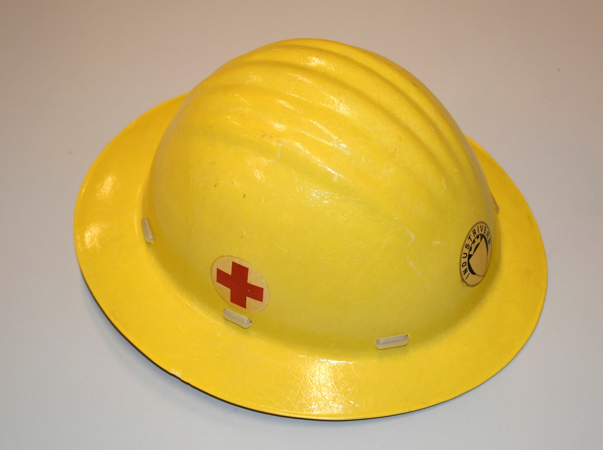 Hjelm i gul plast. Innmaten er laget av plast. Med Industrivern-logo og rødt kors.