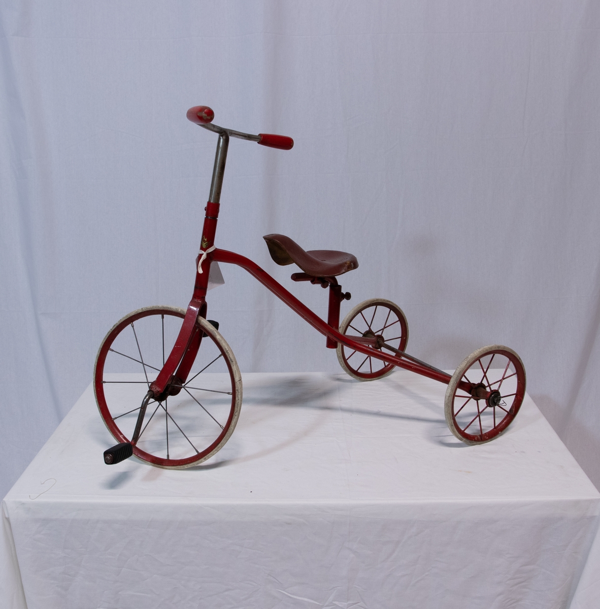 Trehjulsykkel m/rødmalt sykkelramme i stål. To mindre bakhjul, et stort forhjul. Eiker i lettmetall. Hard gråhvit gummi på hjulene. På pedalene tredd to gummi fotstykker, riflemønster. På styret
to sylindriske trehåndtak, rødmalt.