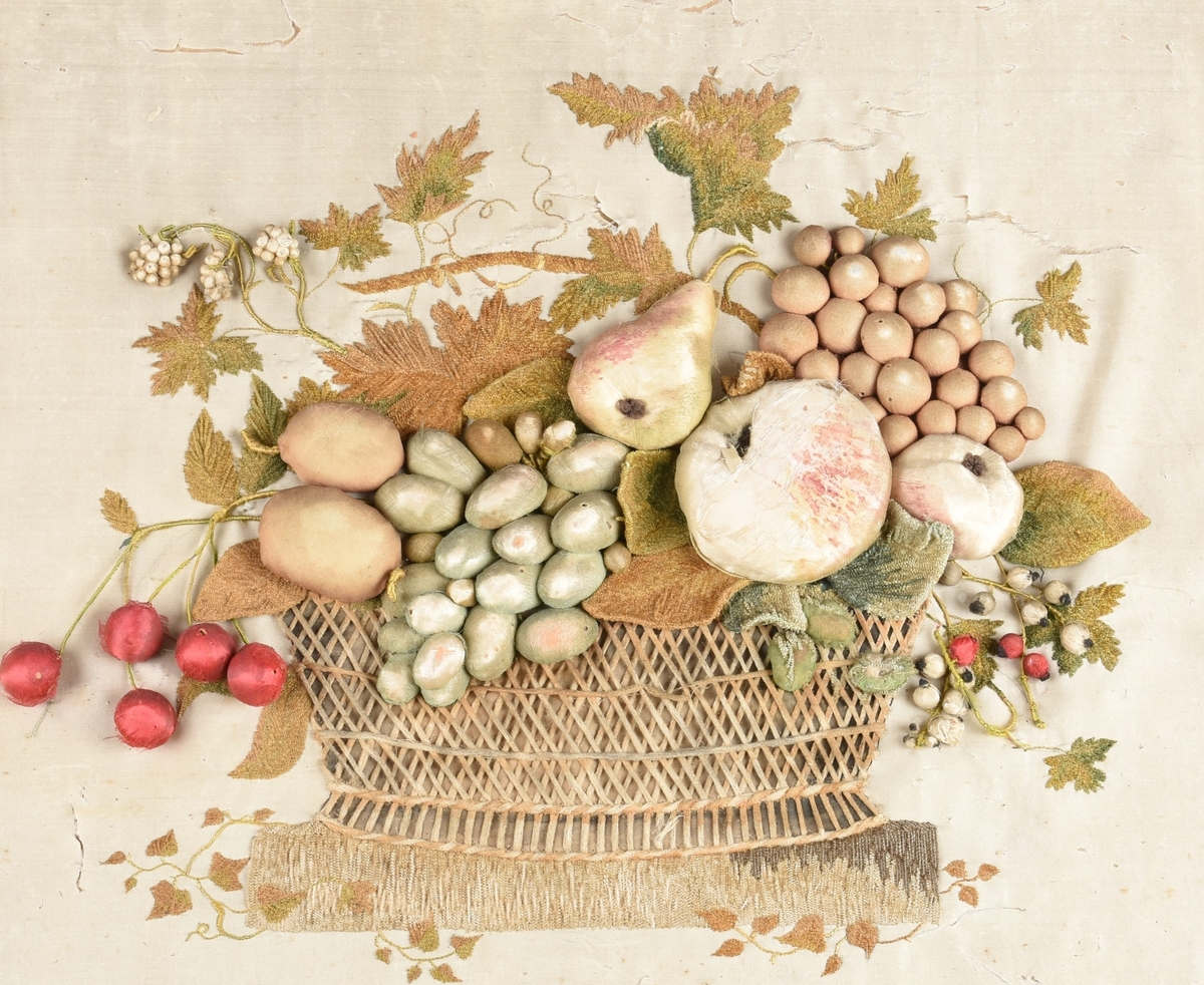 Tekstilbilde. Kvadratisk form. Stilleben med druer, epler, pære, kirsebær m.m. oppi en kurv. Bladornamentikk rundt. Hvit bakgrunn.