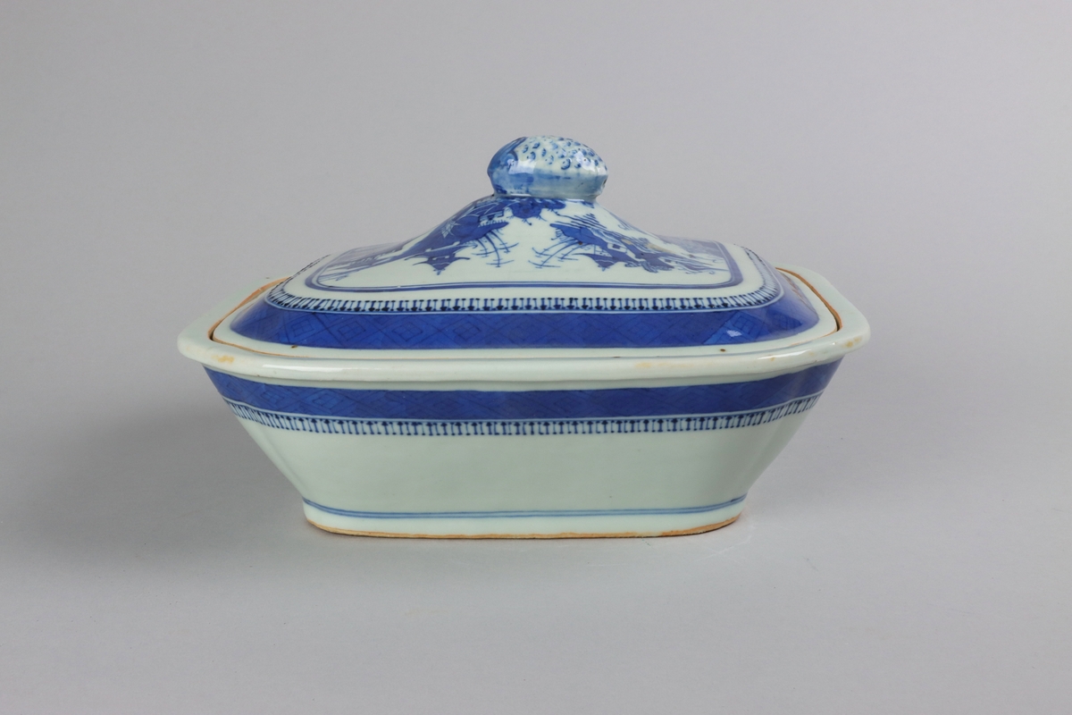 Square form with chamfered corners. The lid with a slightly domed form, on top a knob in form of a fruit. On the lid landscape scenes of pagodas, buildings,  bridges, figures with parasolls, gardens and waters. The edge is decorated with a wide dark blue border with a criss cross pattern. The sides of the dish are below the rim decorated with a criss cross patterned bord. All decor in blue underglaze. The base is not decorated.