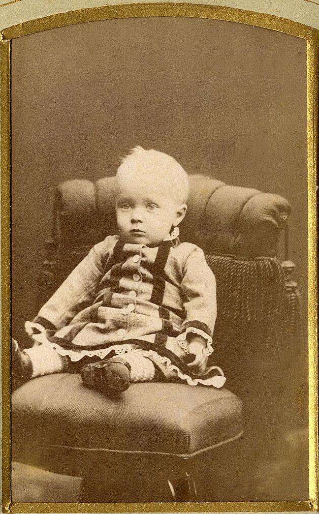 En liten pojke (?) i kolt. Han sitter i en fåtölj hos fotografen. 
Helfigur, halvprofil. Ateljéfoto.
