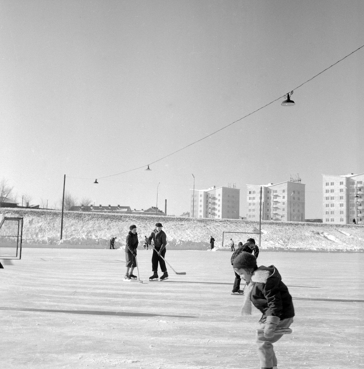 Serie bilder från Lingvallen i Linköping vintern 1955. Anläggningen skapades i början av decenniet, inte minst genom stora, frivilliga insatser av medlemmar i idrottsföreningen Gottfridsbergs IF. Tillkomsten av Lingvallen innebar bland annat att föreningen återupptog bandy på sitt program. I bakgrunden färdigställts punkthusen utmed Åbylundsgatan.