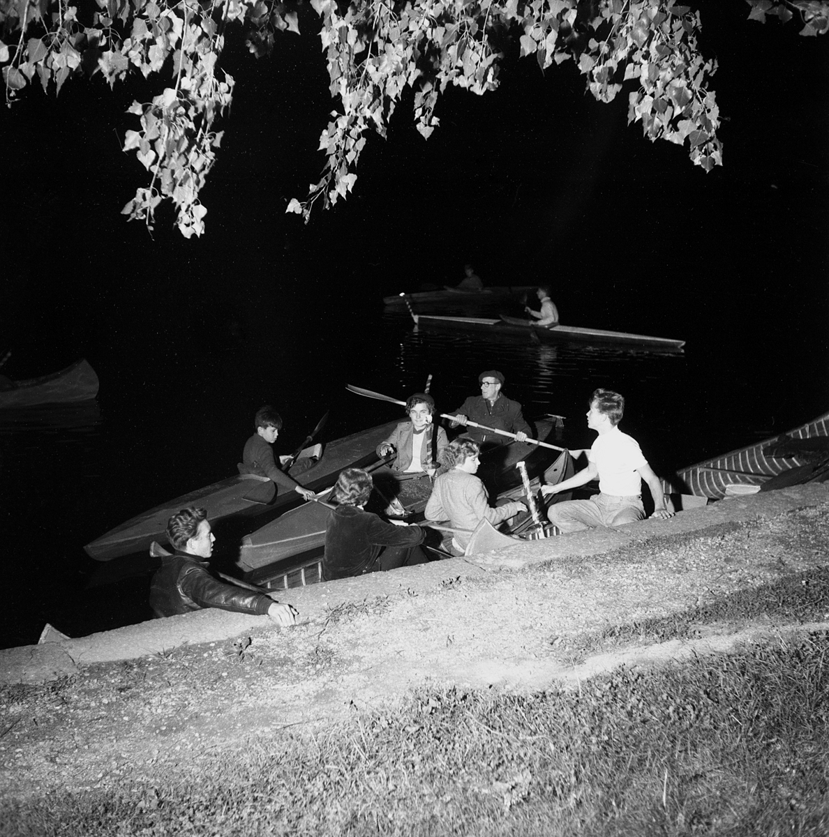 Medlemmar i Linköpings Kanotklubb förbereder ett fackeltåg på Stångåns vatten. Fotografens register ger oss inga detaljer kring arrangemanget men det skedde i september 1954.