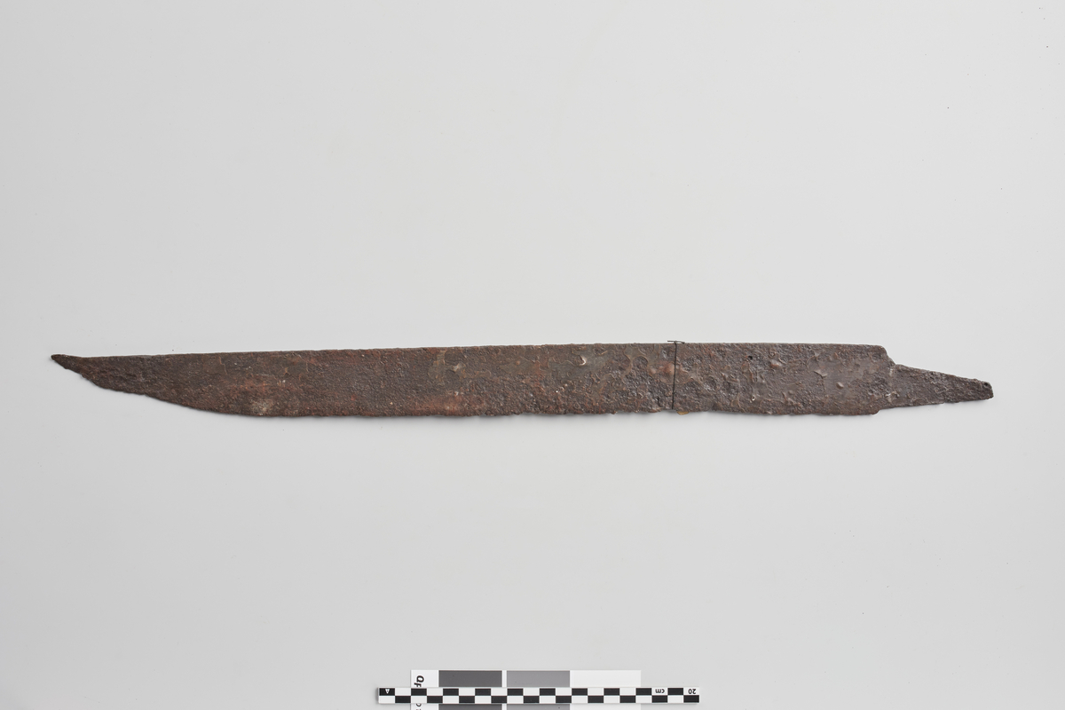 Kort enegget sverd av jern omtr. som R. 499, men med kortere tange. Glødeskall. Lengde 59 cm, derav tangen 7 cm, klingens bredde 4,5 cm.