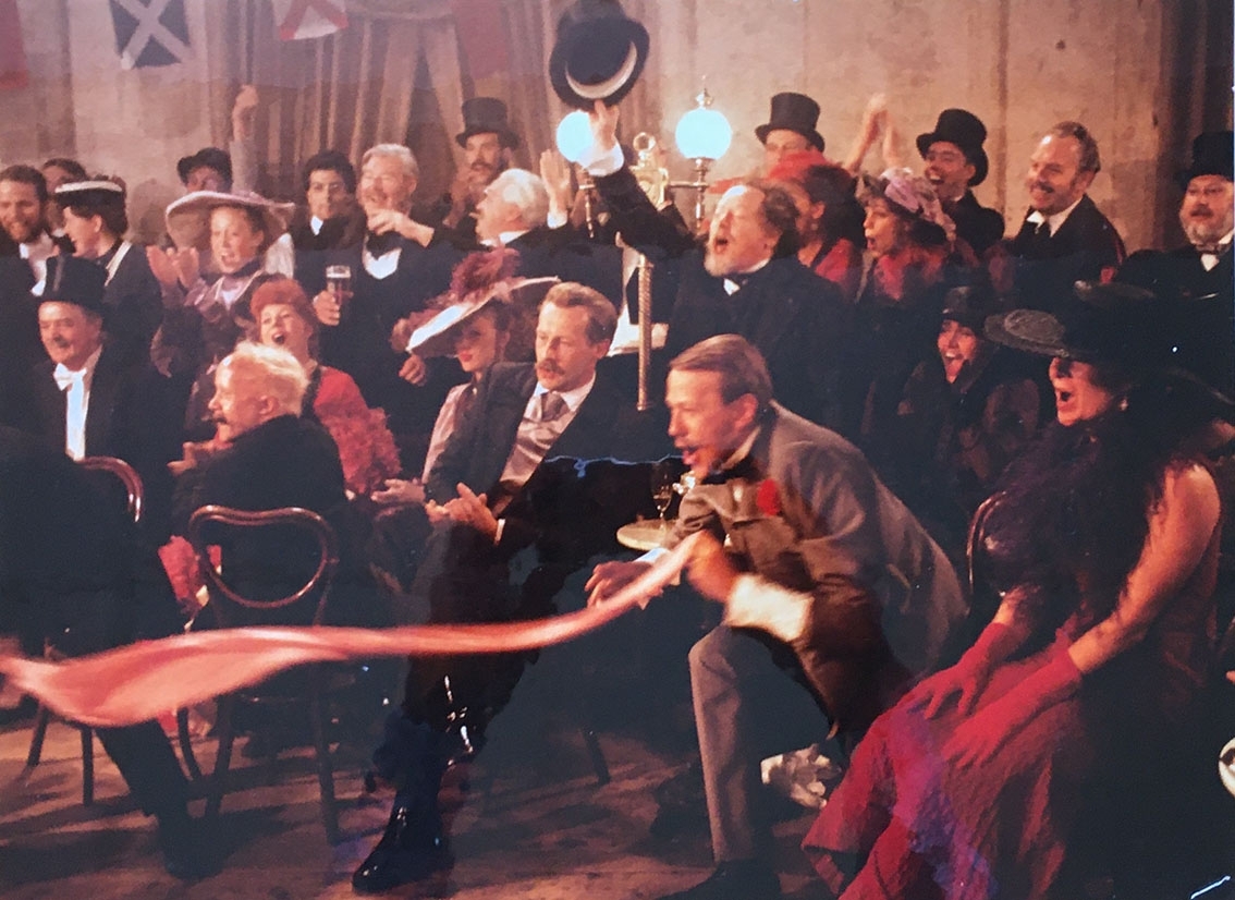 Ett större sällskap män och kvinnor klädda för sent 18900-tal. De står och sitter, tjoar, skriker, applåder och viftar med höga hattar vända mot en can-can-dans utanför bild.