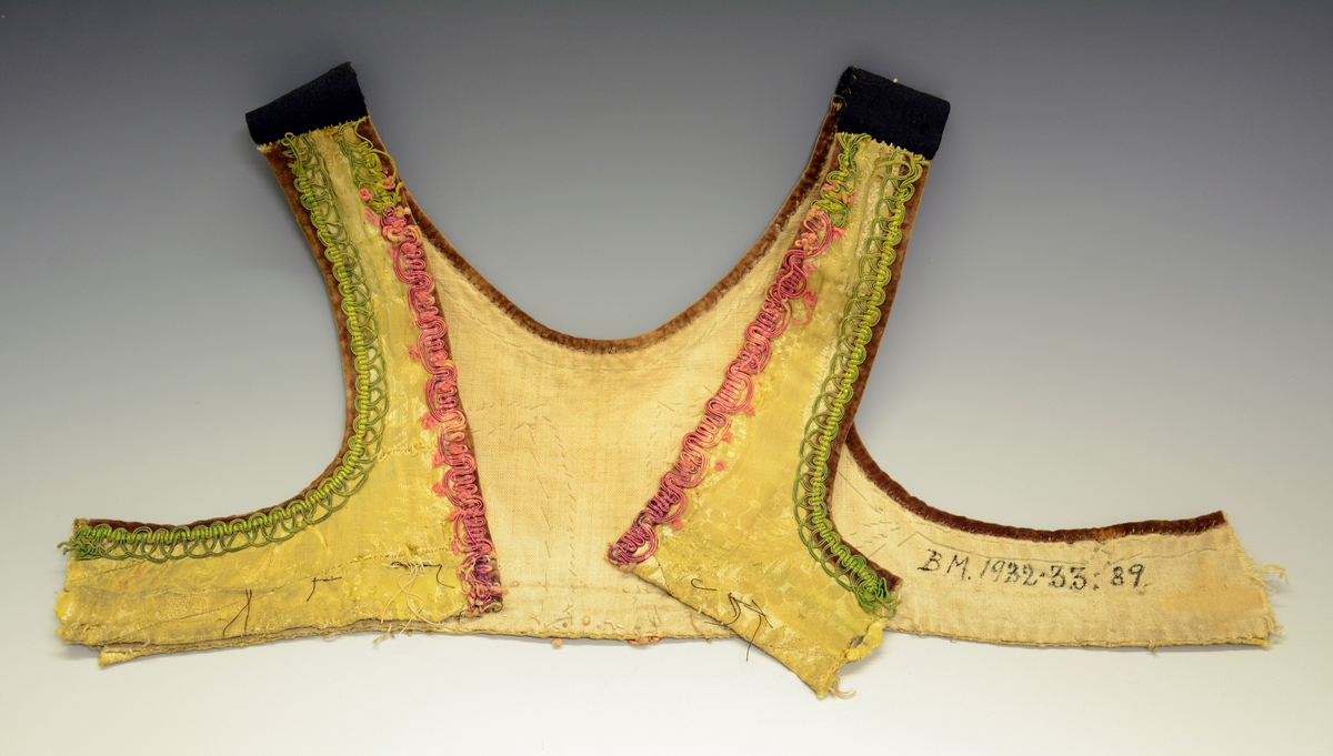 Ovaliv (upplut) av gul silkedamask med bordur av raude og grøne agramanur, Brun fløyelskant og lereftsfor. Ved ermstaupet er livet avklypt.  Fra protokoll.