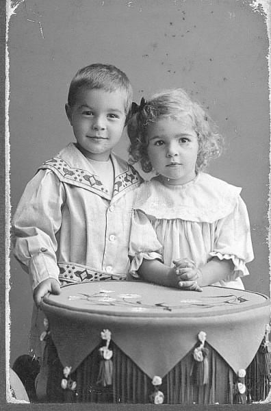 Ateljéfoto av syskonen Greta och Håkan Pehrson, barn till handelsman Oscar Pehrson i Varberg. De står vid ett runt bord med textil beklädnad. Håkan bär en kolt med brodyr på skärp och krage.
