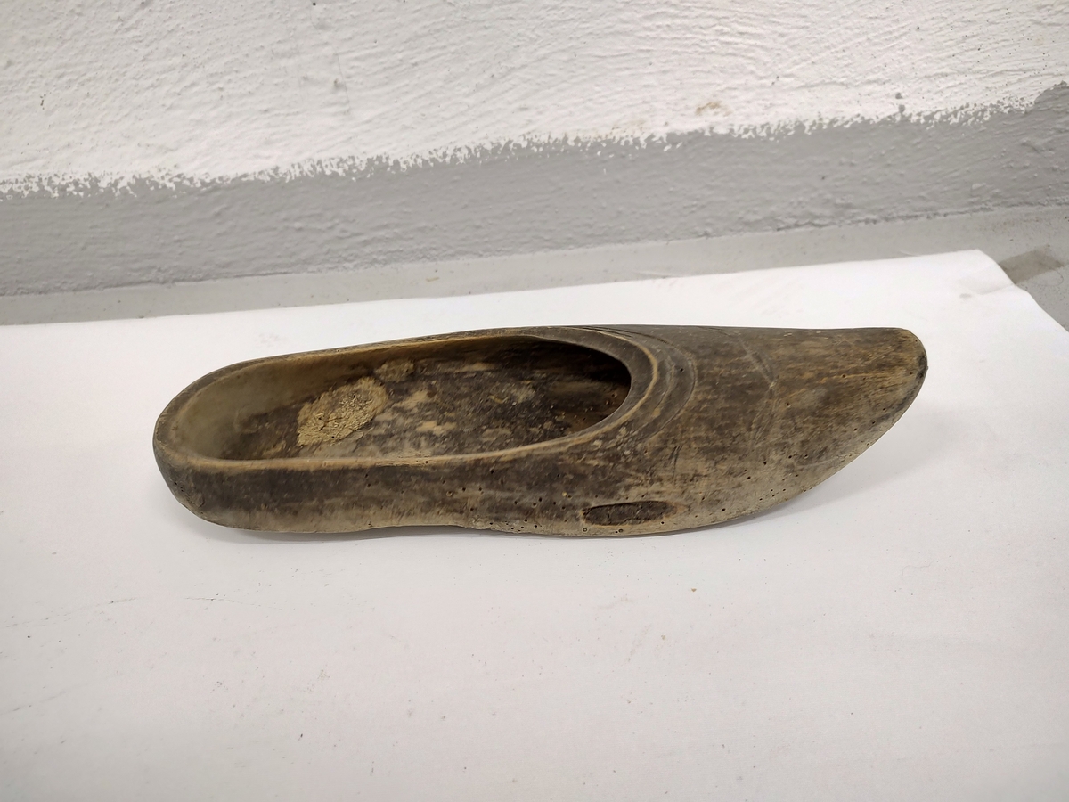 Tresko uten hæl, skåret til og hult ut av et trestykke. Brunmalt. Det er skåret inn et dekorativ mønster langs kanten skoen.