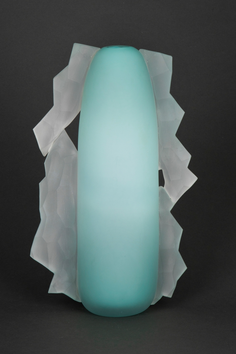 Vase med høyt ovalt korpus. Vasen er bred nederst og blir smalere mot toppen. Vasen har en smal munning. Turkisblå farge på korpus. På to av sidene er det tilført noen abstrakte former som går ned langs hele korpus. Disse er lagd i slipt, hvitt opalt glass.