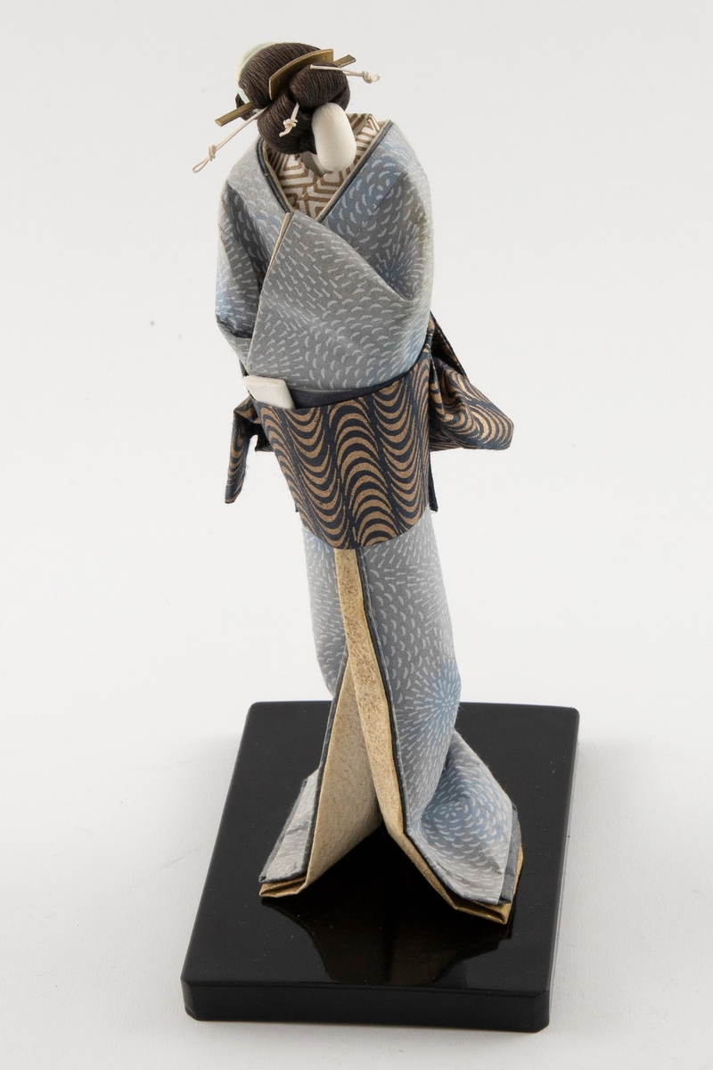 Håndlaget dukke i japansk bretteteknikk. Alle deler er laget i papir. En trepinne er festet i dukken og til en svartmalt tresokkel. Dukken har oppsatt hår og er ikledd en lyseblå kimono med hvitt mønster, en mønsteret innerkimono og mønsteret obi i blå- og bruntoner.