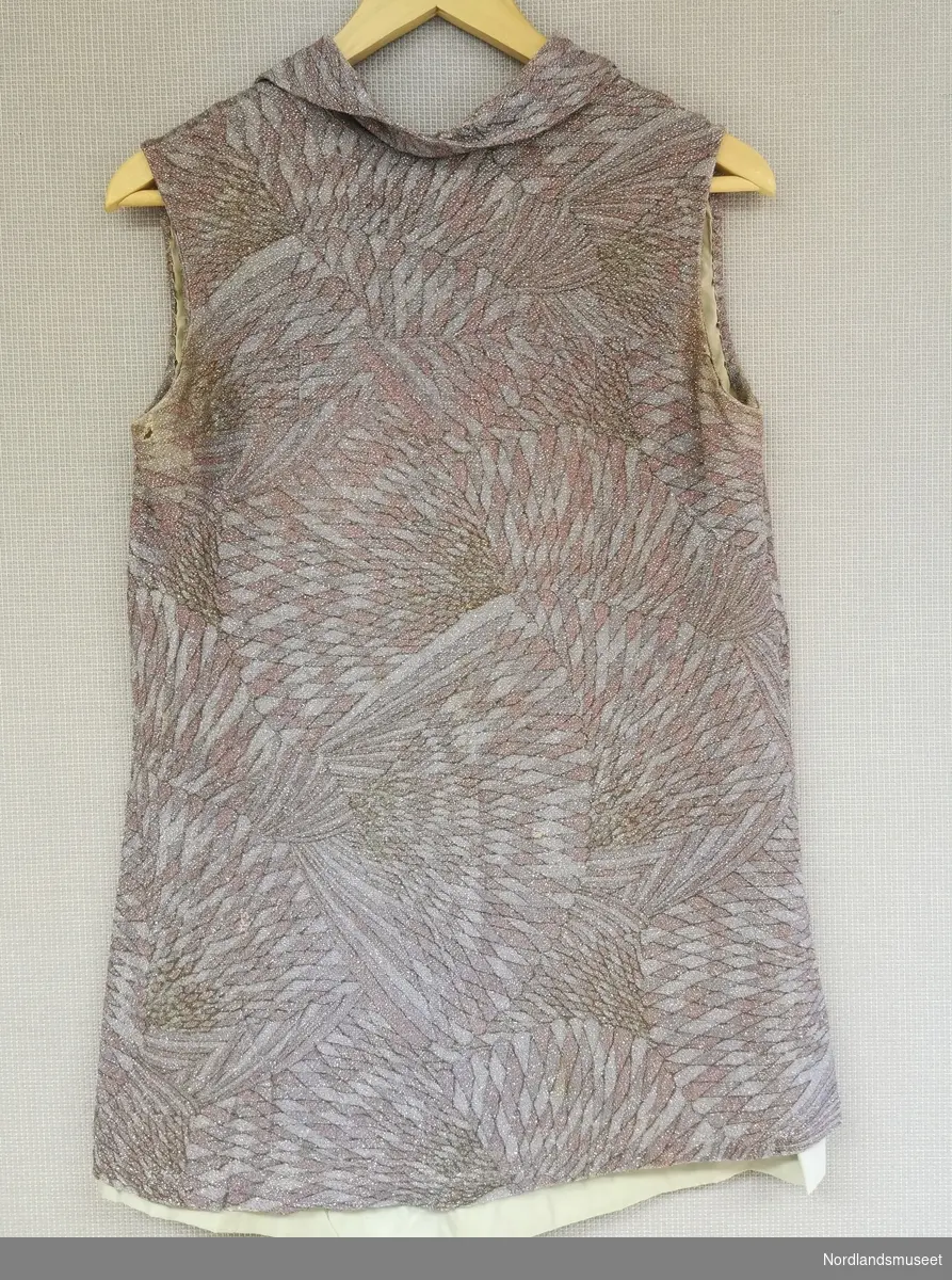 Ermeløs kjole med krage og knappelukking bak i halsen. Kjolen består av to lag, hvor det ytterste laget er mønstret og laget av tekstil med glitter i.