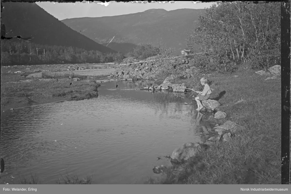 Gutt med hjemmelaget fiskestang sittende ved elvebredd i Månaelven. Kvitåi synes i fjellsiden. Bolighus på Svaddesiden nedenfor gutten. Kjøreledning på jernbanesporet langs elva.