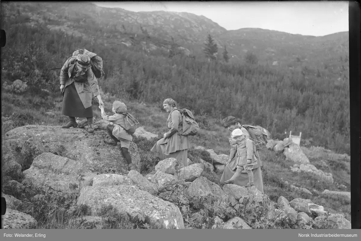 Tre kvinner og to barn på fjelltur. Alle bærer sekk på ryggen. En av kvinnene hjelper gutten opp på en stein. Kvinnene er kledd i skjørt og kåpe med hodeplagg. Barna i knickers og kåpe med hodeplagg.