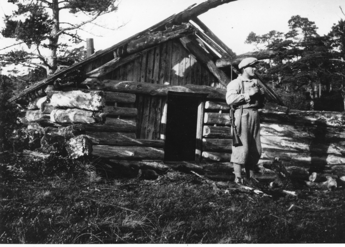 Hans Eilif Fredriksen Strøm med jaktutstyr står utenfor en enkel jakthytte, Rundslettbua, Strøm, Melandsjø, Hitra.