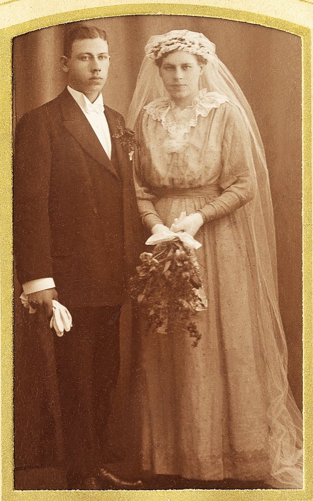 En brudgum i bonjour med brud i färgad brudklänning och brudslöja m.m.
Text under fotot med blyerts: "Ottilia + Theodor Svensson".
Helfigur. Ateljéfoto.