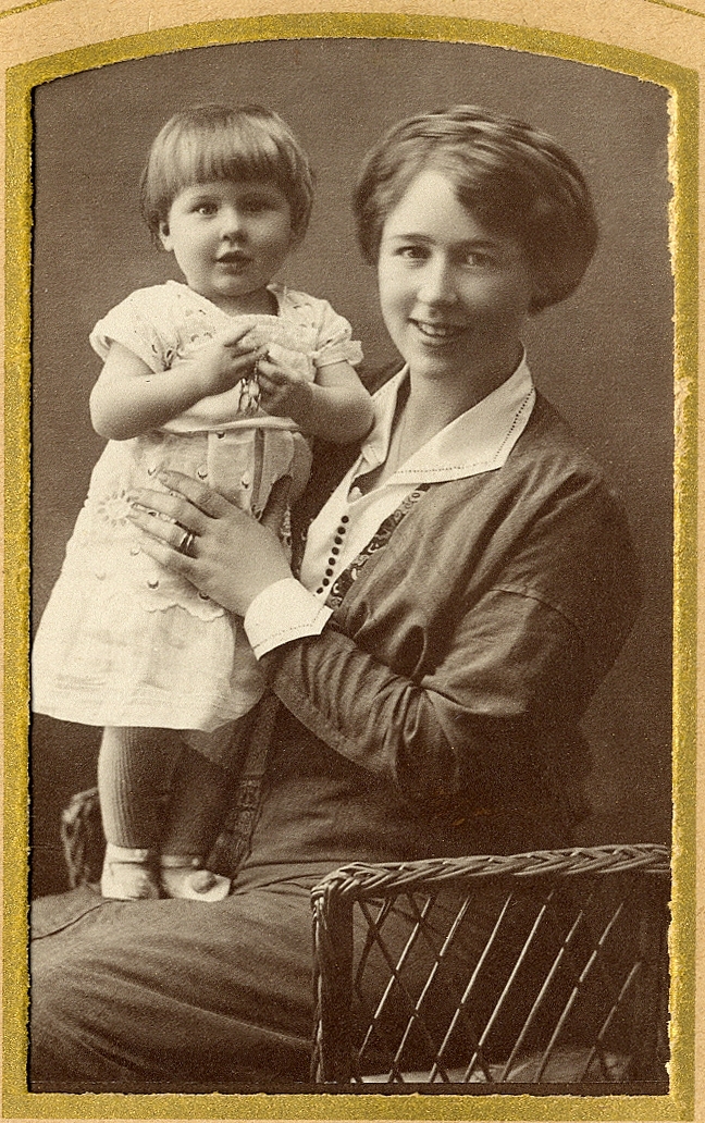 En kvinna i klänning med vit krage sitter i en korgstol och håller om en liten flicka i ljus klänning.
Under fotot text med blyerts: "Arbetsgivare i Eksjö 1915". På baksidan står skrivet: "Ann-Mari 15 månader".
Knäbild. Ateljéfoto.