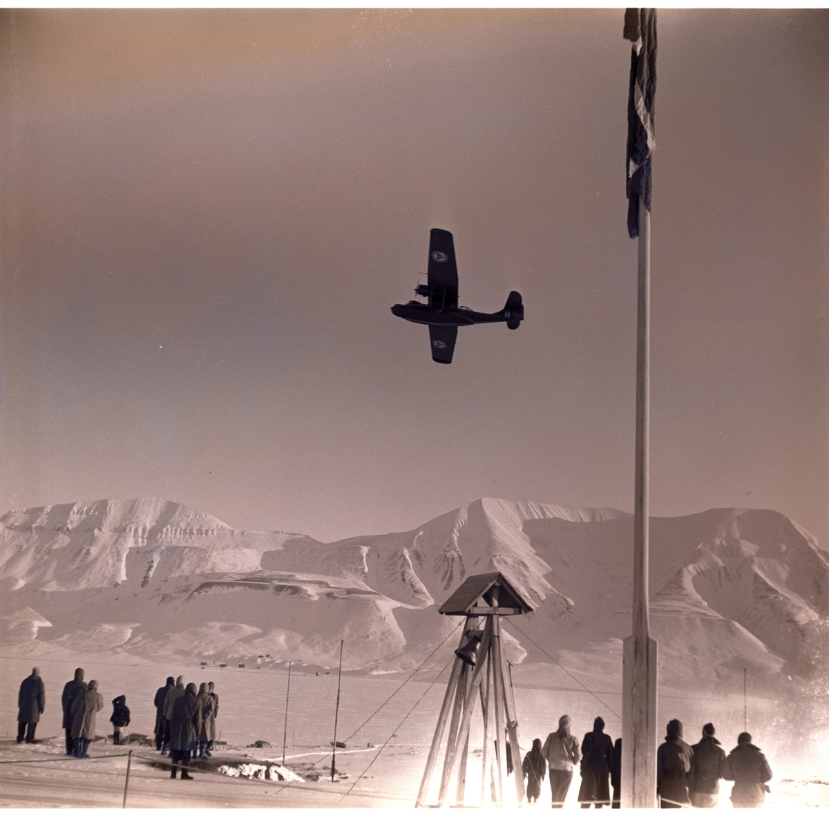 Postflyet dropper postsekker over myra i Longyearbyen 18.mars 1956. Folk som følger med fra bautaen på Skjæringa.
