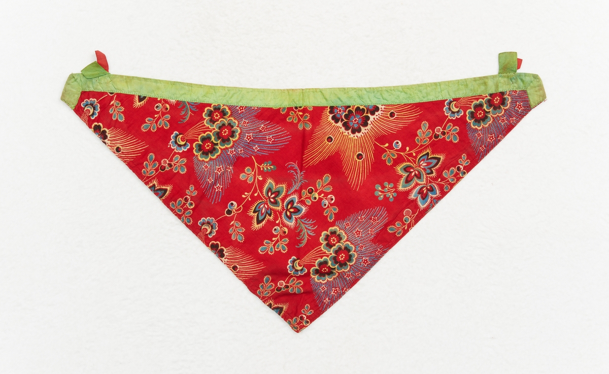 Halskläde, astrasu till Rättviks kvinnliga sockendräkt. Trekantigt av kattun med tryckt mönster i vitt, gult, svart, blått och grönt på röd botten. Foder av bomullslärft med tryckta små röda och gröna blommor i rader. Den något bågformade långsidan är kantad med ett ljusgrönt sidenband, en påsydd tamp av samma gröna band och under den en röd fastsydd på vardera sidan, för fastsättning av brosch.