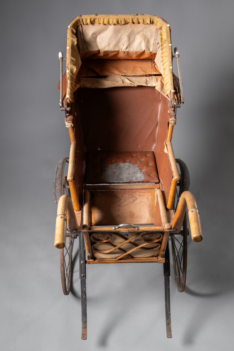 Barnvagn, kombinerad ligg- och sittvagn. Fotlådan går att fälla upp/ner till liggande respektive sittande läge. Stomme av järn bestående av underrede med två stora framhjul och två små bakhjul, samt två stödben (så att vagnen inte tippar fram). Hjulen är av trä med plåtskoning och ekrar av järn. Korg av trä, betsat, dekorerad utvändigt med böjd rotting. Invändigt fodrad med brunt konstläder. Stoppad sits. Två höga, böjda trähandtag. Sufflett av brunt konstläder på metallställning. Suffletten går att fälla upp och ner samt går att ta av. Handtag av vitt porslin. Runt sufflettens ytterkant är ett textilt band med udd fäst. Bandet har ursprungligen varit fäst med nubb, senare fäst med häftstift (de två översta på höger sida i samband med registreringen). Band av textil runt kanten under suffletten.  Sits och fotlådan nötta, skador på suffletten.